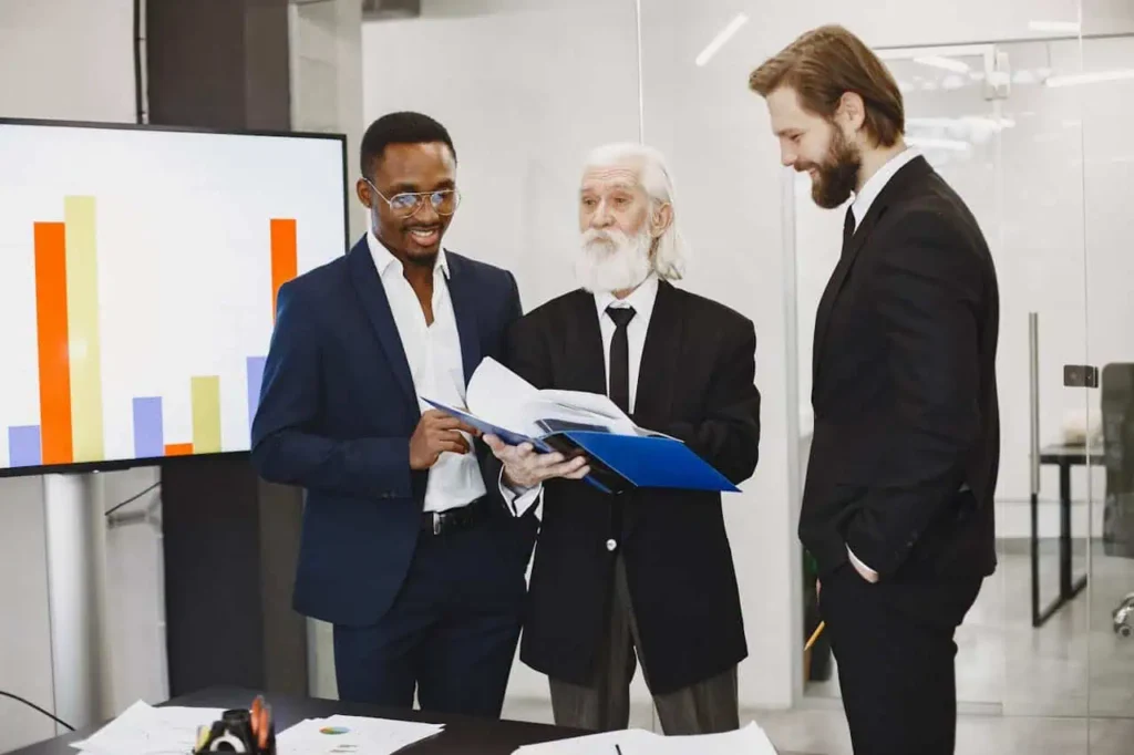 Three businessmen reviewing documents together beside large bar chart screen.