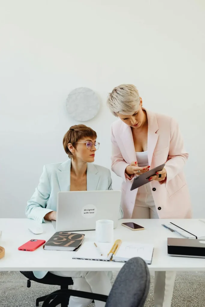 Two businesswomen collaborating while using tablet at modern white desk.